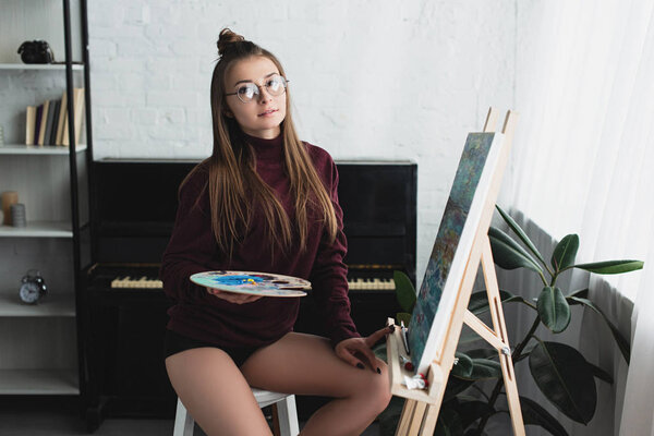 girl in burgundy sweater sitting and looking at camera while painting at home 