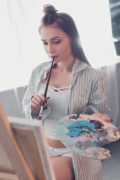 attractive artist holding palette, biting paintbrush and looking at easel in living room