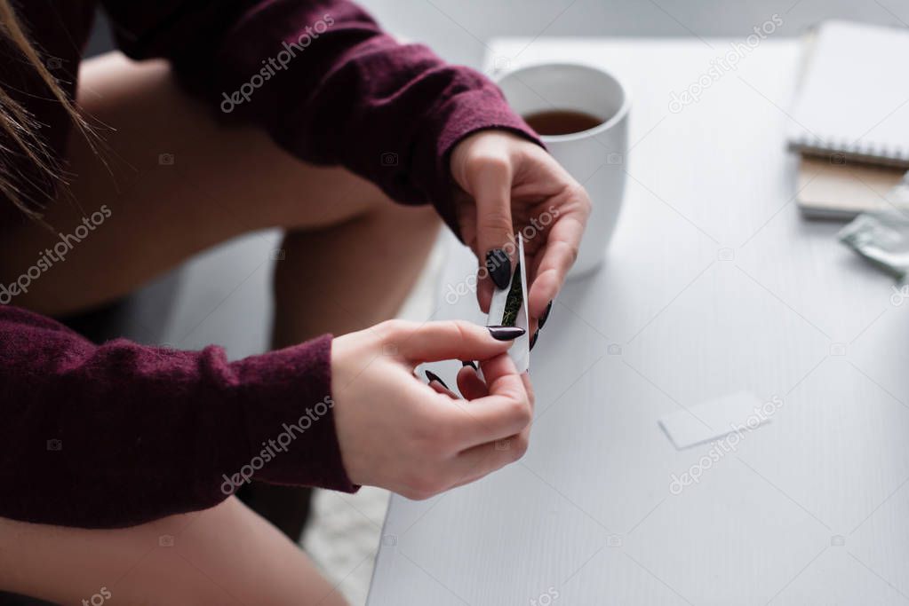 cropped view of girl sitting and rolling marijuana joint at home 