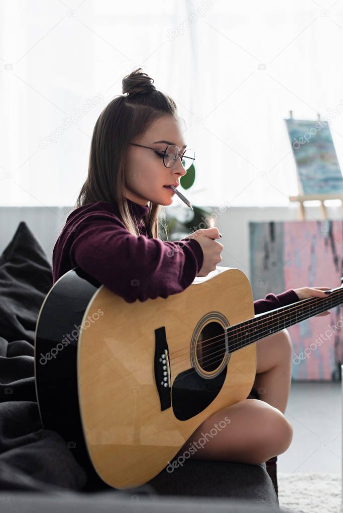 attractive girl sitting on couch, lighting marijuana joint and holding guitar at home