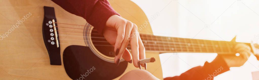 cropped view of girl sitting, holding marijuana joint and playing guitar at home