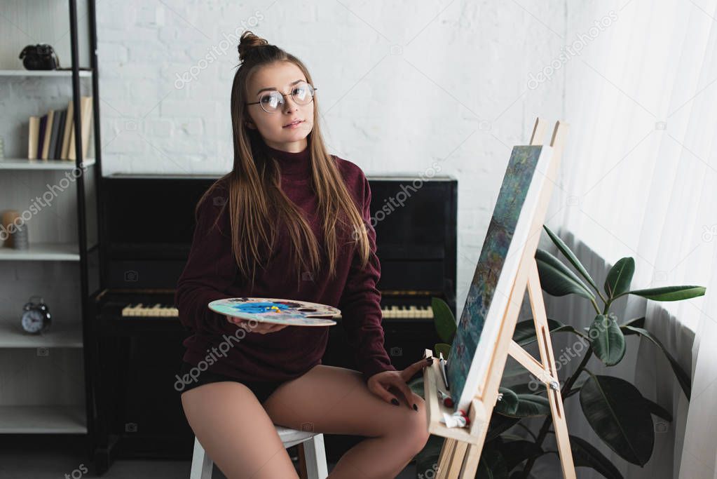 girl in burgundy sweater sitting and looking at camera while painting at home 