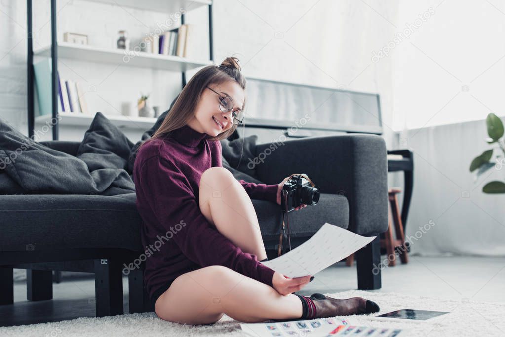 attractive photographer sitting on carpet near sofa, holding film camera and looking at photos in living room