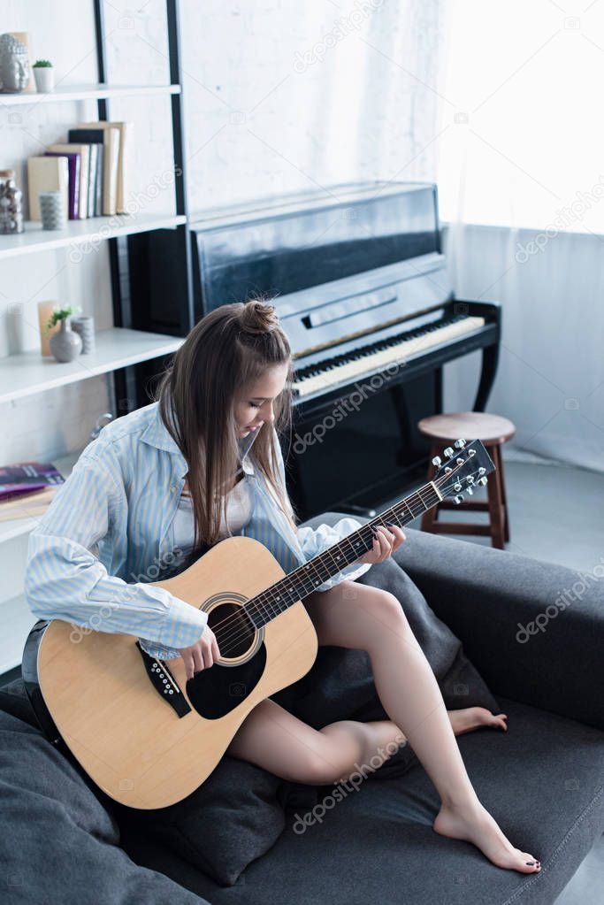 attractive musician sitting on couch and playing acoustic guitar in living room