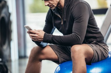 fitness topu üzerinde oturan ve spor salonunda smartphone kullanarak sporcu görüntü kırpılmış 