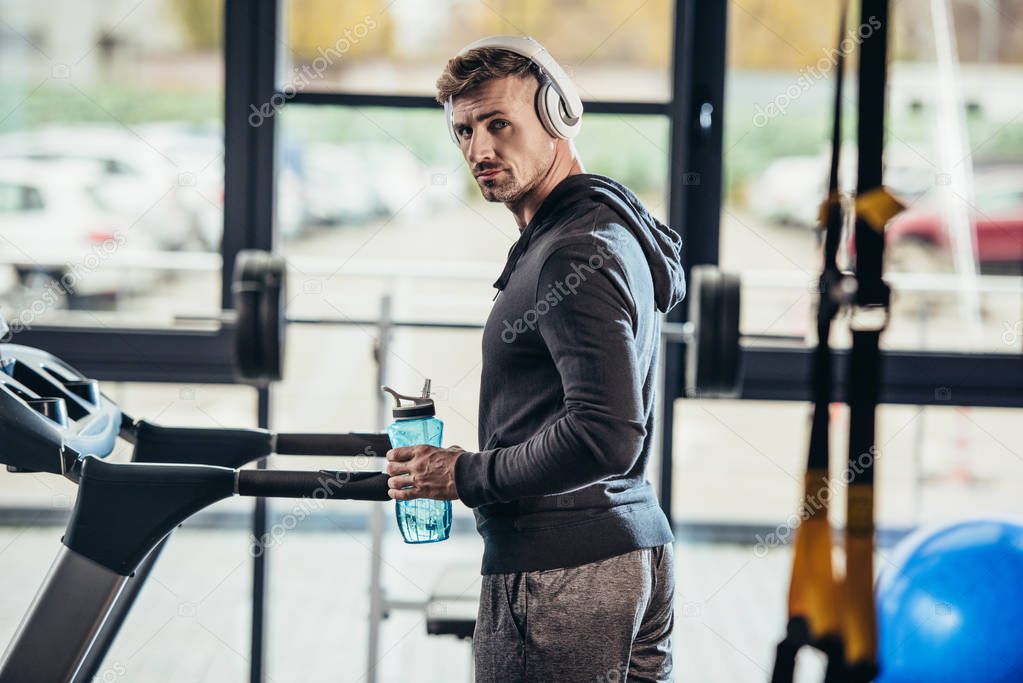 portrait of handsome sportsman in hoodie standing near treadmill with sport bottle in gym and looking at camera