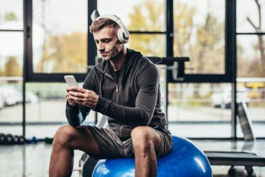 Smartphone kullanarak ve fitness topu spor salonunda otururken yakışıklı sporcu 