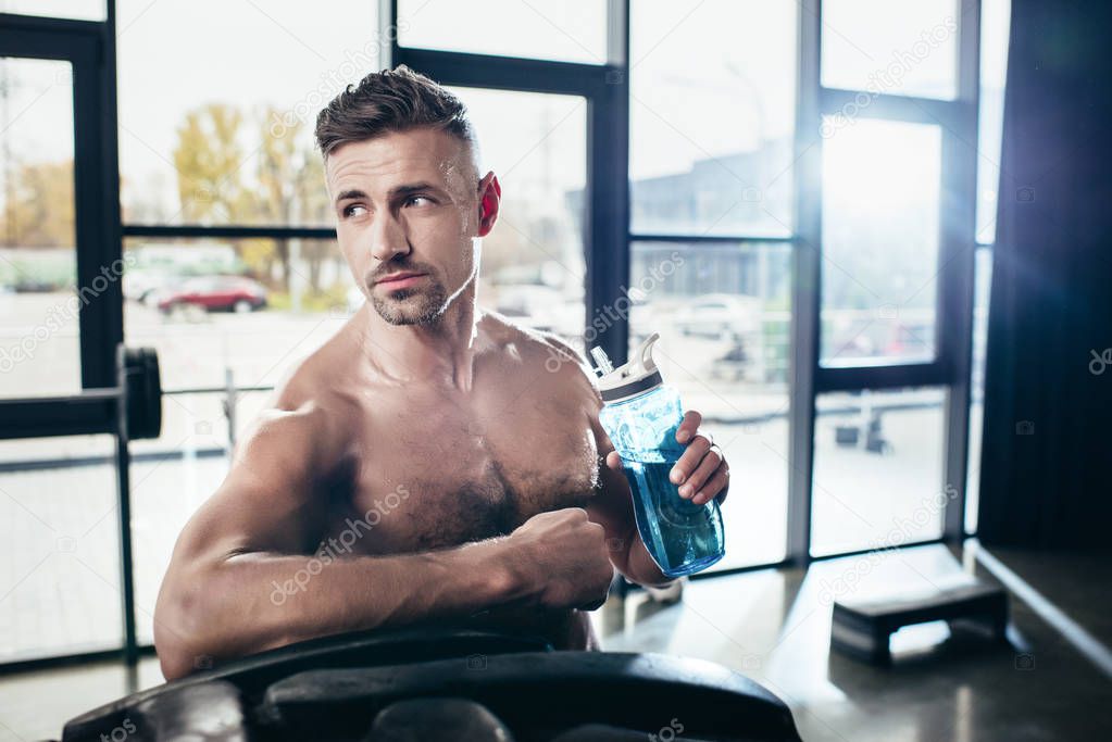 handsome shirtless sportsman leaning on tire and holding sport bottle in gym, looking away