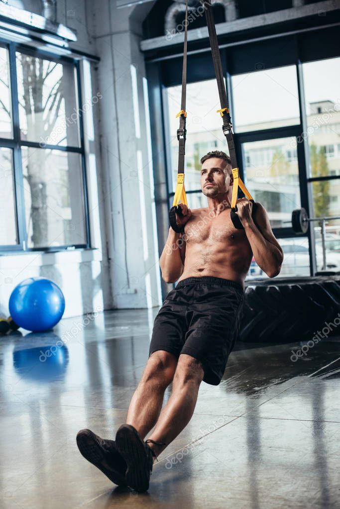 handsome shirtless sportsman training with suspension straps in gym 