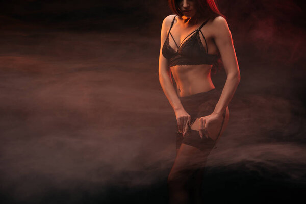 обрезанный вид молодой женщины в эротическом белье позирует в темном дымчатом помещении
