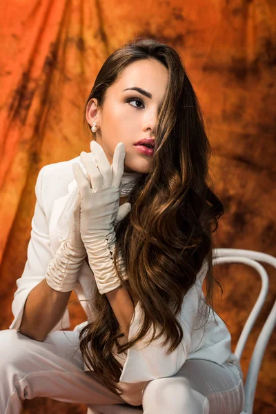 beautiful girl with long hair in white suit and gloves sitting on chair on textured background