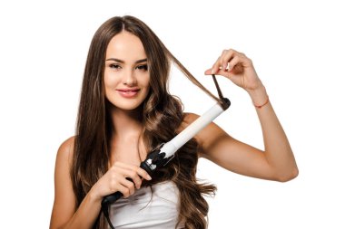 smiling girl with long brown hair using curling iron and looking at camera isolated on white clipart