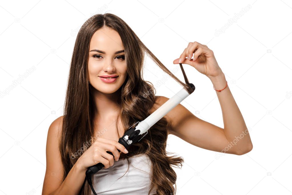 smiling girl with long brown hair using curling iron and looking at camera isolated on white