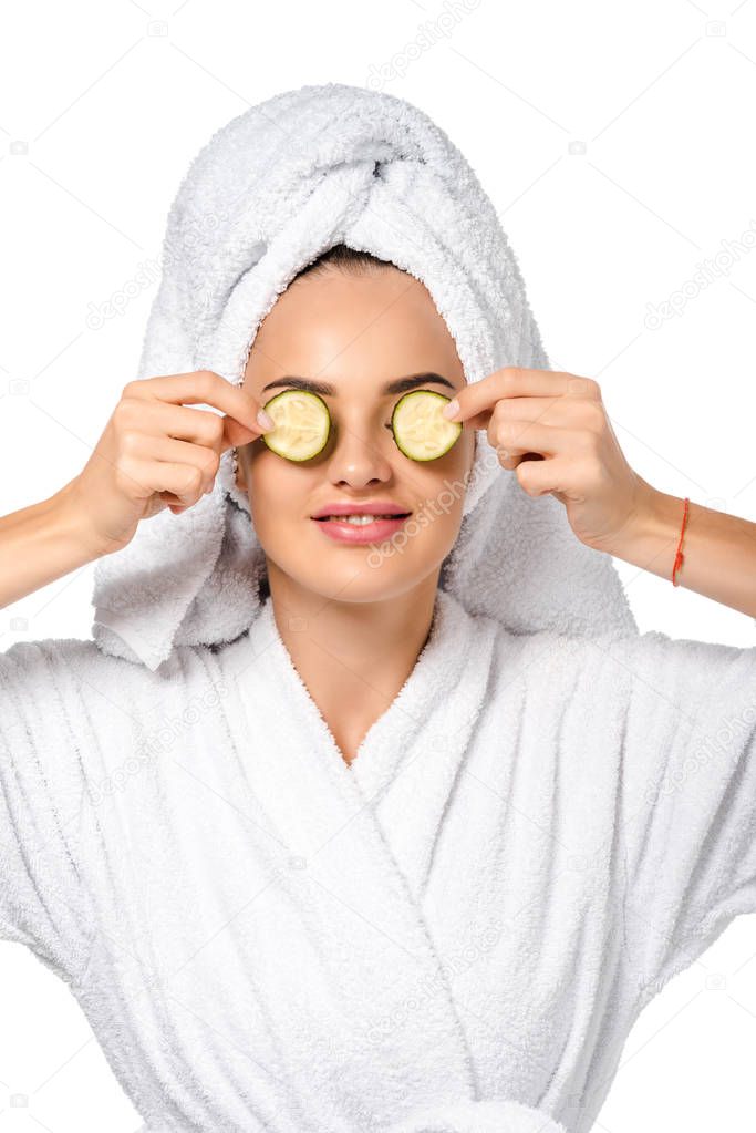 beautiful girl in bathrobe holding cucumber slices near eyes and smiling isolated on white