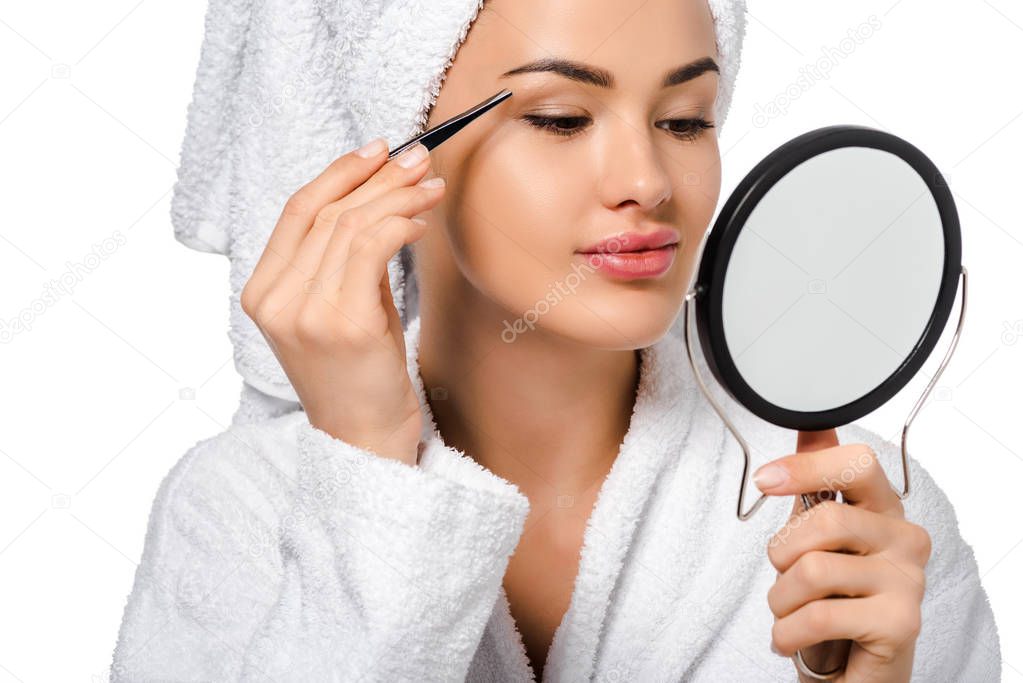 close up view of beautiful girl in bathrobe looking at mirror and tweezing eyebrows isolated on white
