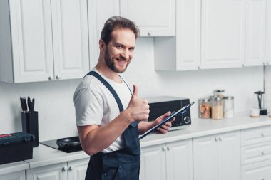 yakışıklı yetişkin sakallı tamirci holding Pano gülümseyen ve mutfak hareketle yukarı yapıyor 