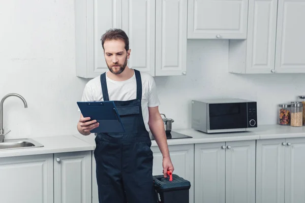 Serious Bearded Repairman Working Overall Holding Toolbox Looking Clipboard Kitchen — Stock Photo, Image