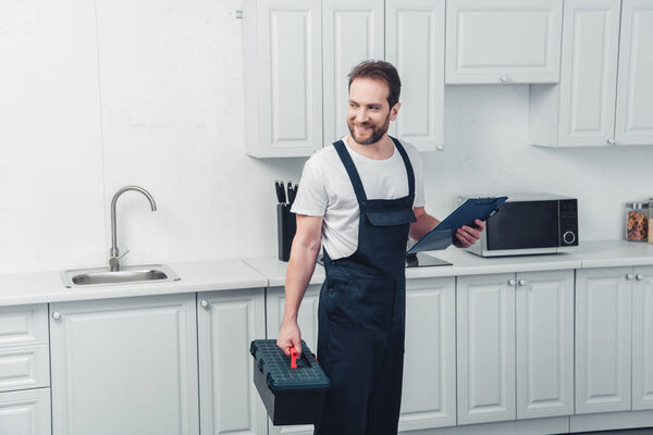 cheerful adult repairman in working overall holding toolbox and clipboard in kitchen at home