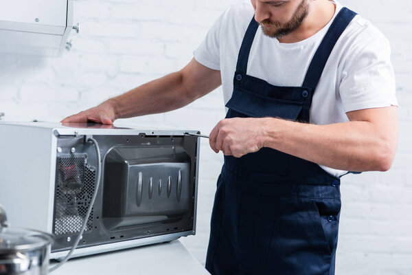 partial view of bearded handyman repairing microwave oven by screwdriver in kitchen
