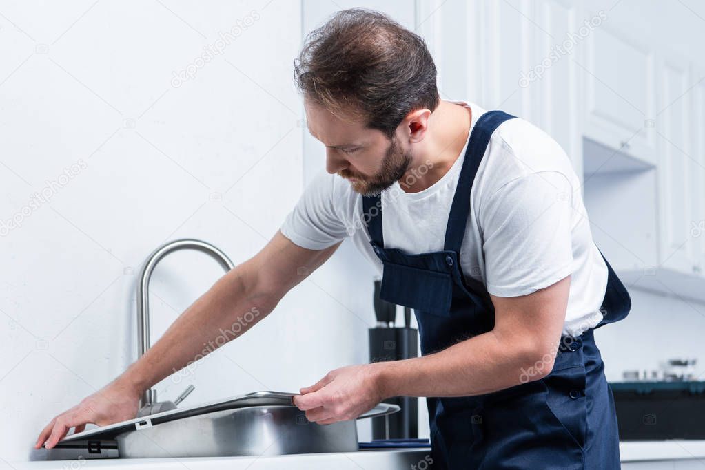 adult bearded repairman in working overall fixing sink in kitchen 