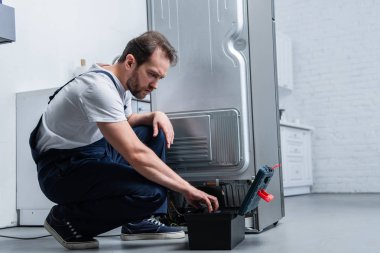 repairman in working overall near taking tools from toolbox broken refrigerator in kitchen