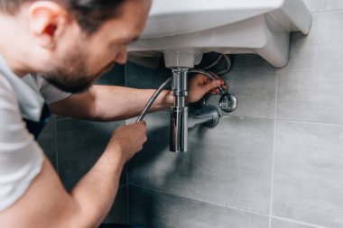 selective focus of male plumber in working overall fixing sink in bathroom  clipart