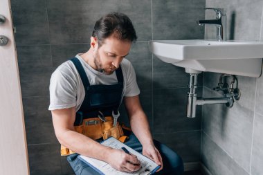 Pano kırık lavabo banyo yakınındaki yazma toolbelt ile yetişkin erkek tesisatçı