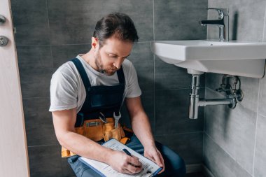 adult male plumber with toolbelt writing in clipboard near broken sink in bathroom clipart