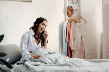 Yatağın üstüne oturan ve smartphone tarafından konuşmak güzel gülümseyen genç kadın