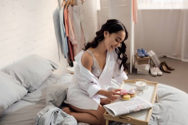 çekici gülümseyen genç kadının yatak odasında kahvaltı yaparken macaron çerez holding yüksek açılı görünüş