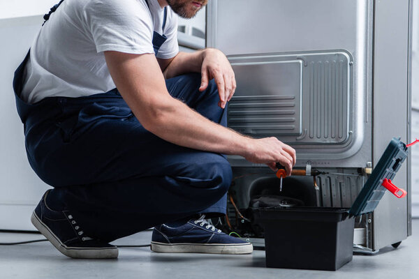 cropped shot of craftsman in working overall near taking screwdriver from toolbox broken refrigerator in kitchen