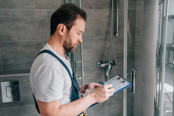 クリップボードに浴室でシャワーを確認しながらノートを作る男性配管工の側面図 — ストック写真