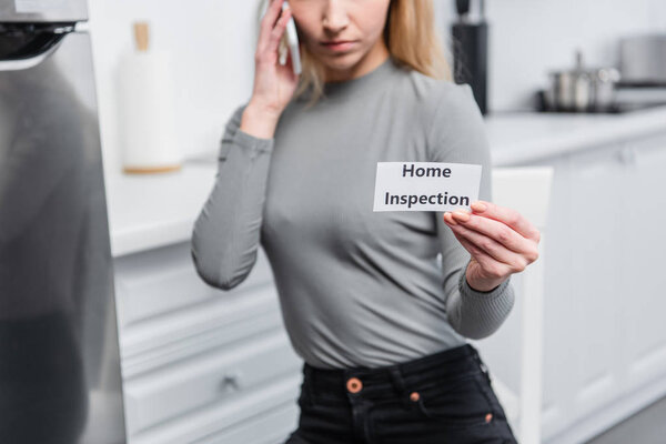 partial view of young woman holding card with lettering home inspection and talking on smartphone in kitchen 