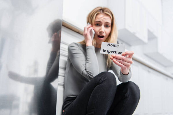shocked young woman holding card with lettering home inspection and talking on smartphone near broken refrigerator in kitchen 