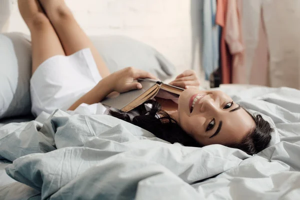 本とカメラに笑顔でベッドに横たわって幸せな若いアジア女性 — ストック写真