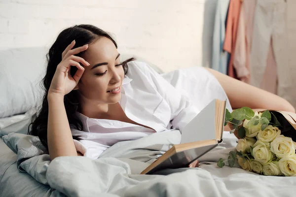 Красива Усміхнена Азіатська Дівчина Лежить Ліжку Букетом Троянд Читає Книгу — стокове фото