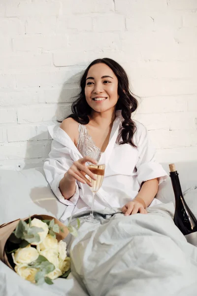 幸せなアジア美人ベッドでシャンパン グラスを保持ブラと白いシャツで — ストック写真