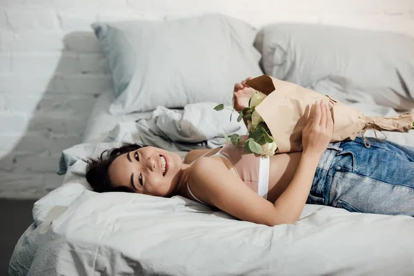 幸せな若い女の美しい花の花束を持って ベッドの上に横たわっている間カメラで笑顔 — ストック写真
