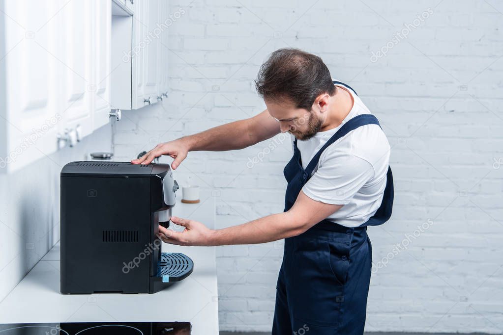 side view of handyman in working overall repairing coffee machine in kitchen