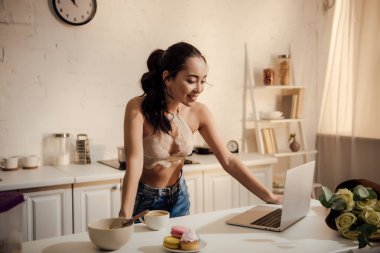 dantel bralette ve evde kahvaltı sırasında dizüstü kullanarak kot pantolon güzel gülümseyen genç kadın 