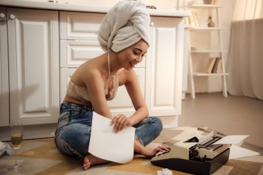 smiling young woman with towel on head holding paper and using typewriter while sitting on floor clipart