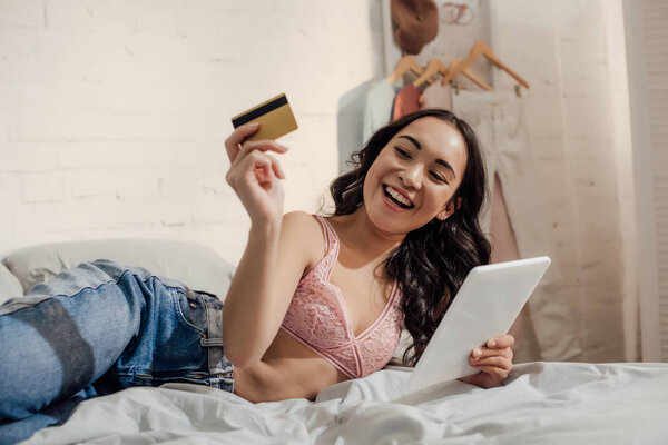 cheerful young woman shopping online with digital tablet and credit card in bedroom 