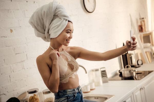 beautiful smiling asian girl with towel on head taking selfie with smartphone in kitchen 