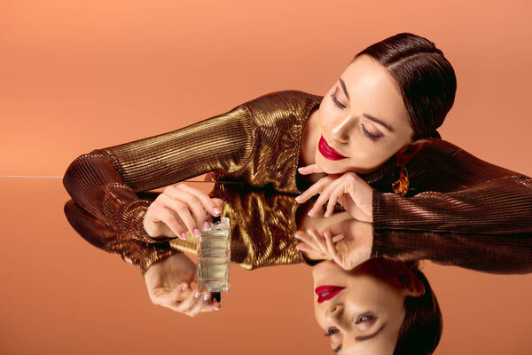 beautiful smiling woman in golden clothes with glamorous makeup, perfume bottle and mirror reflection posing isolated on orange