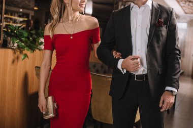 cropped view of boyfriend and girlfriend in red dress walking in restaurant and holding hands clipart