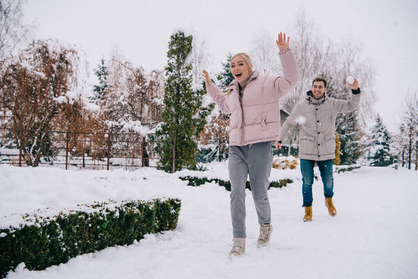 joyful woman running from boyfriend throwing snowball in winter 