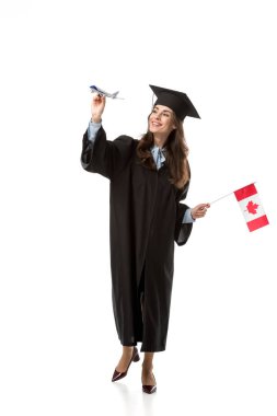 smiling female student in academic gown holding canadian flag and plane model isolated on white, studying abroad concept clipart