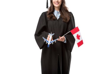 cropped view of female student in academic gown holding canadian flag and plane model isolated on white, studying abroad concept clipart
