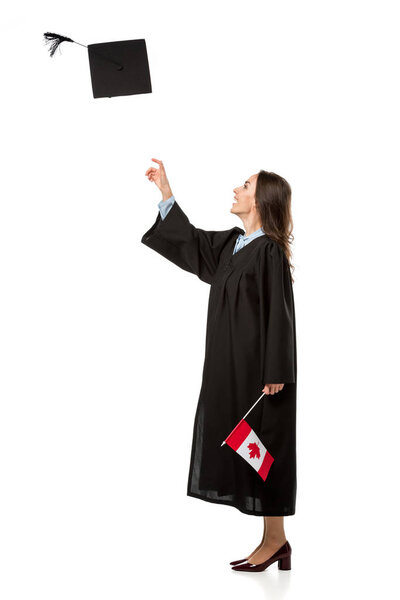 female student in academic gown holding canadian flag and throwing mortarboard in air isolated on white