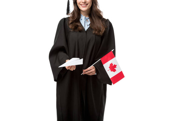 cropped view of female student in academic gown holding canadian flag and paper plane isolated on white